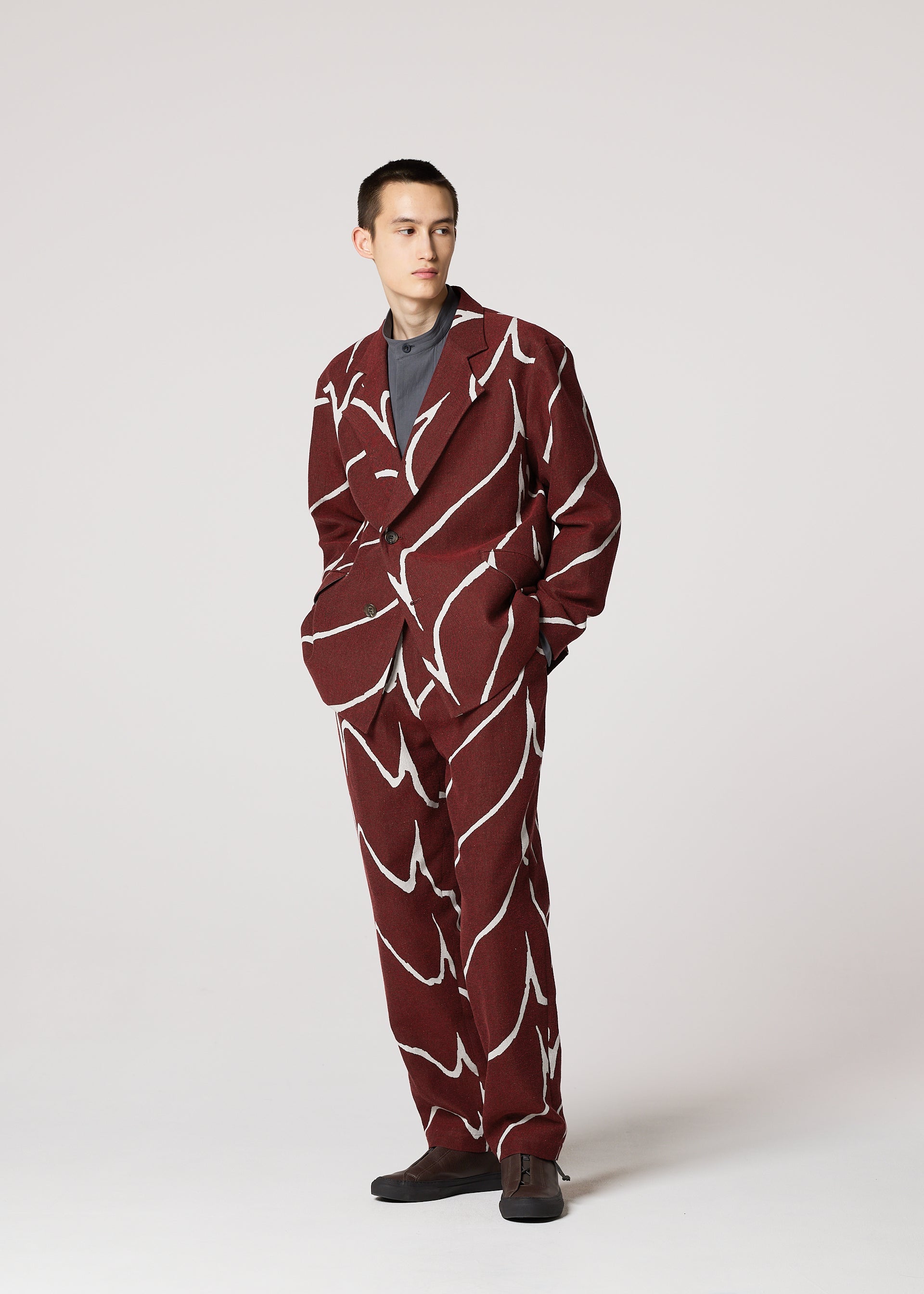 Zoomed view A person stands with hands in pockets, wearing the IM MEN EARTH maroon tailored jacket with a bold jacquard wave pattern, paired with a gray shirt and black shoes, set against a plain light background.
