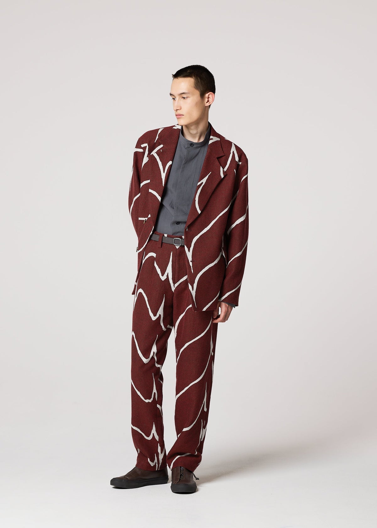 A person models the IM MEN EARTH suit—a maroon jacquard wave pattern with bold white lines—paired with a gray shirt and dark shoes, standing against a plain light background, looking slightly to the side. view 9 of 9