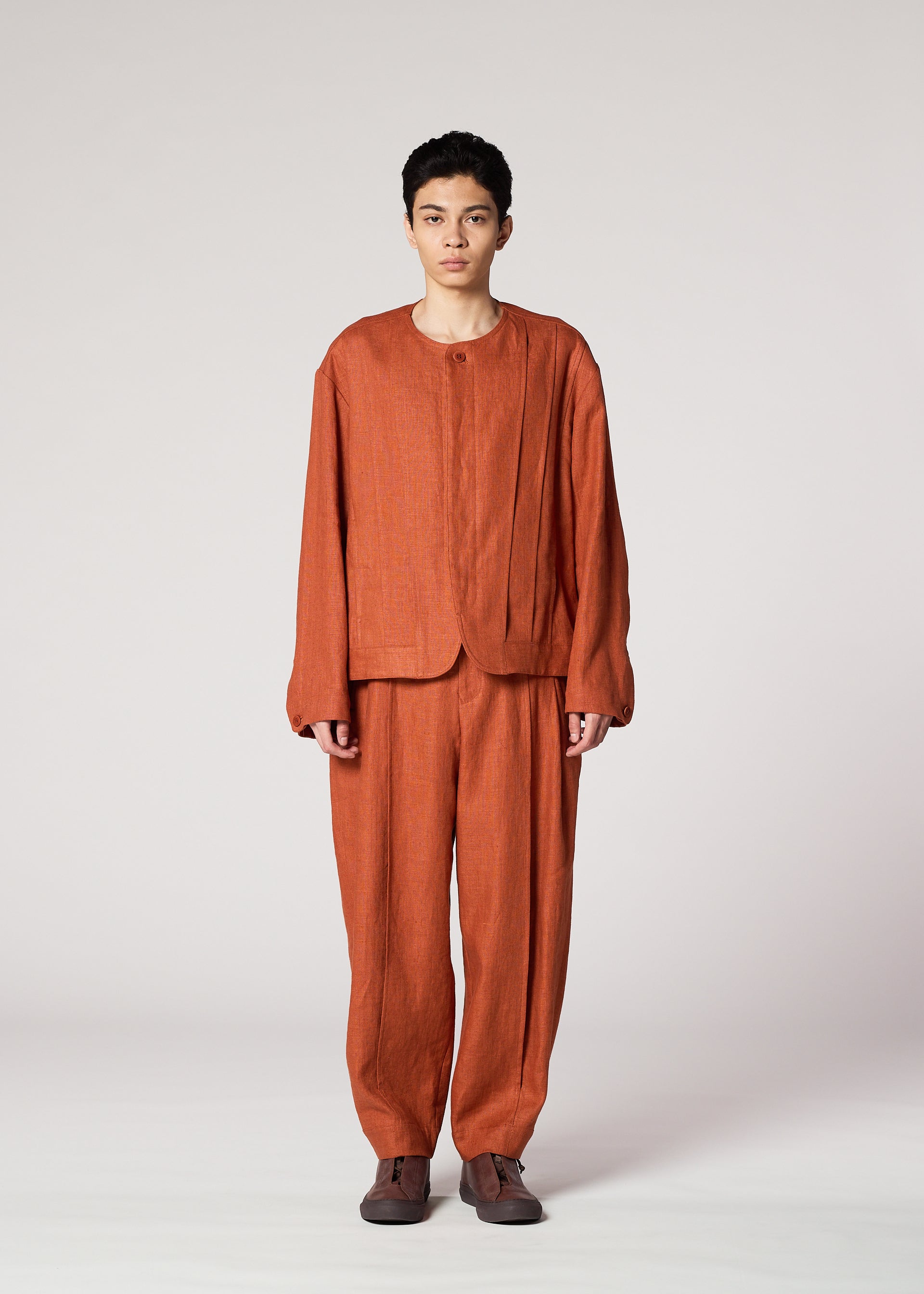 Zoomed view A person faces forward against a plain, light background, wearing IM MEN's TOUGH LINEN: a loose-fitting, rust-colored collarless jacket and matching textured trousers, paired with brown shoes.