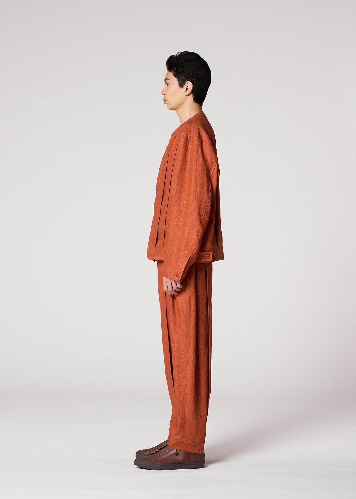 A person stands in profile against a light plain background, wearing IM MEN’s TOUGH LINEN: a loose-fitting, rust-colored collarless jacket and matching pants with natural texture, paired with dark brown shoes. view 7 of 9