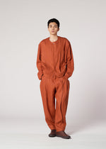 Against a plain light gray background, a person wears IM MEN's TOUGH LINEN set: a rust-colored, loose-fitting, round-neck collarless jacket paired with matching pants and brown shoes.