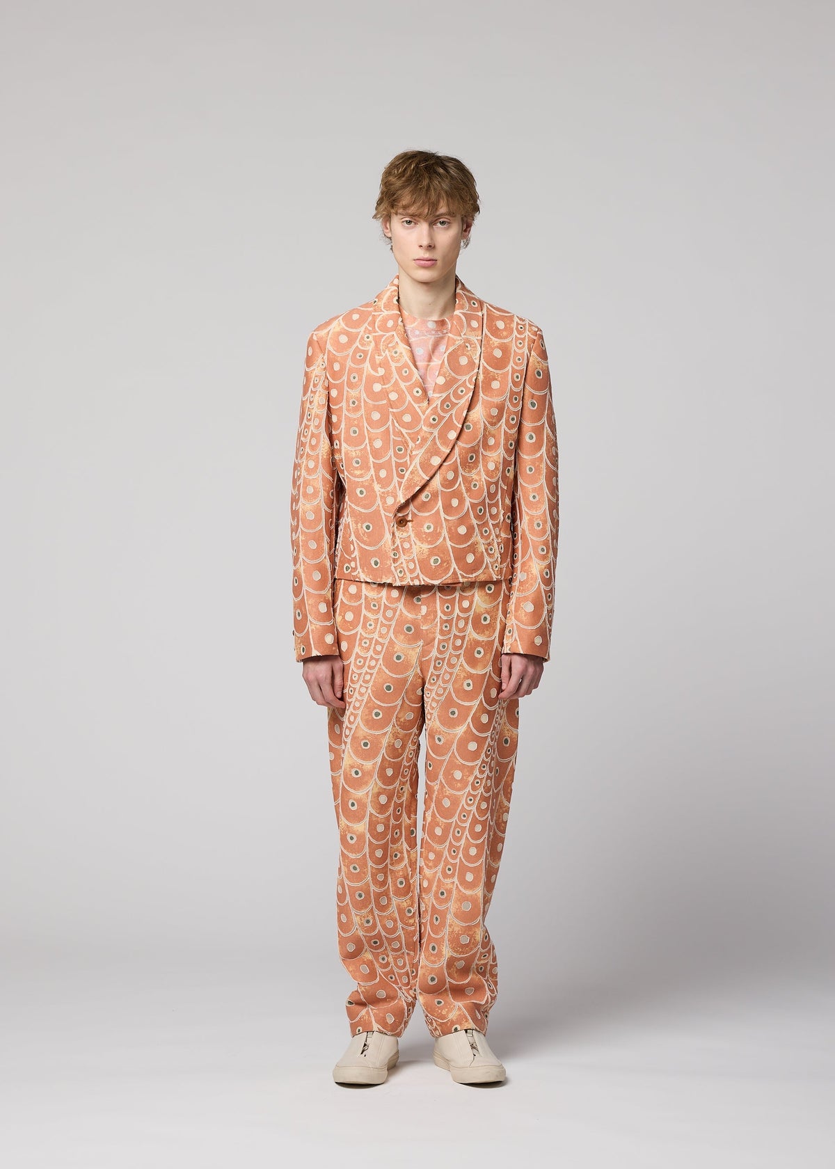 A person wearing the IM MEN UROKOMON peach-colored suit with a white scale-like pattern, white shoes, and a neutral expression stands facing forward against a plain light gray background. view 5 of 8