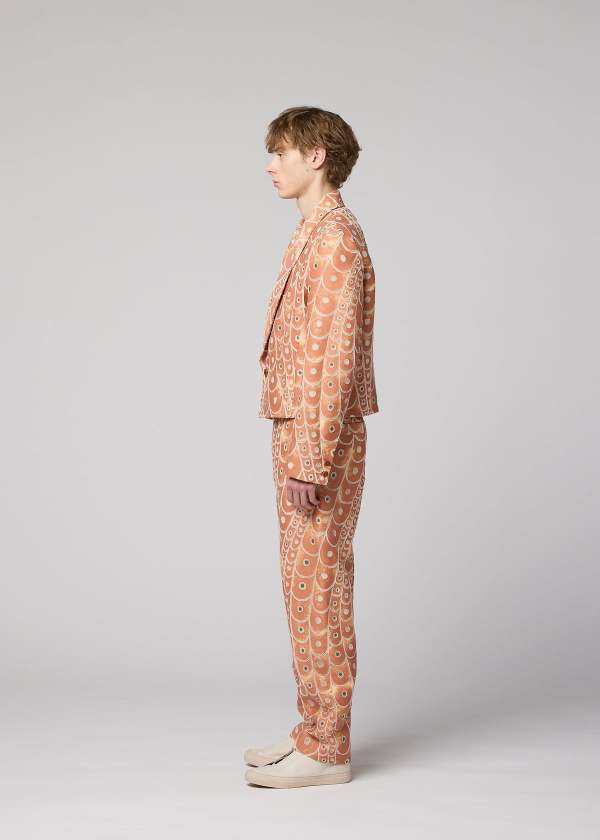 A person stands in profile against a plain light gray background, wearing IM MEN's UROKOMON tan suit with a scale-inspired design by Shoji Kamoda, featuring a short jacket and paired with cream-colored shoes. view 6 of 8