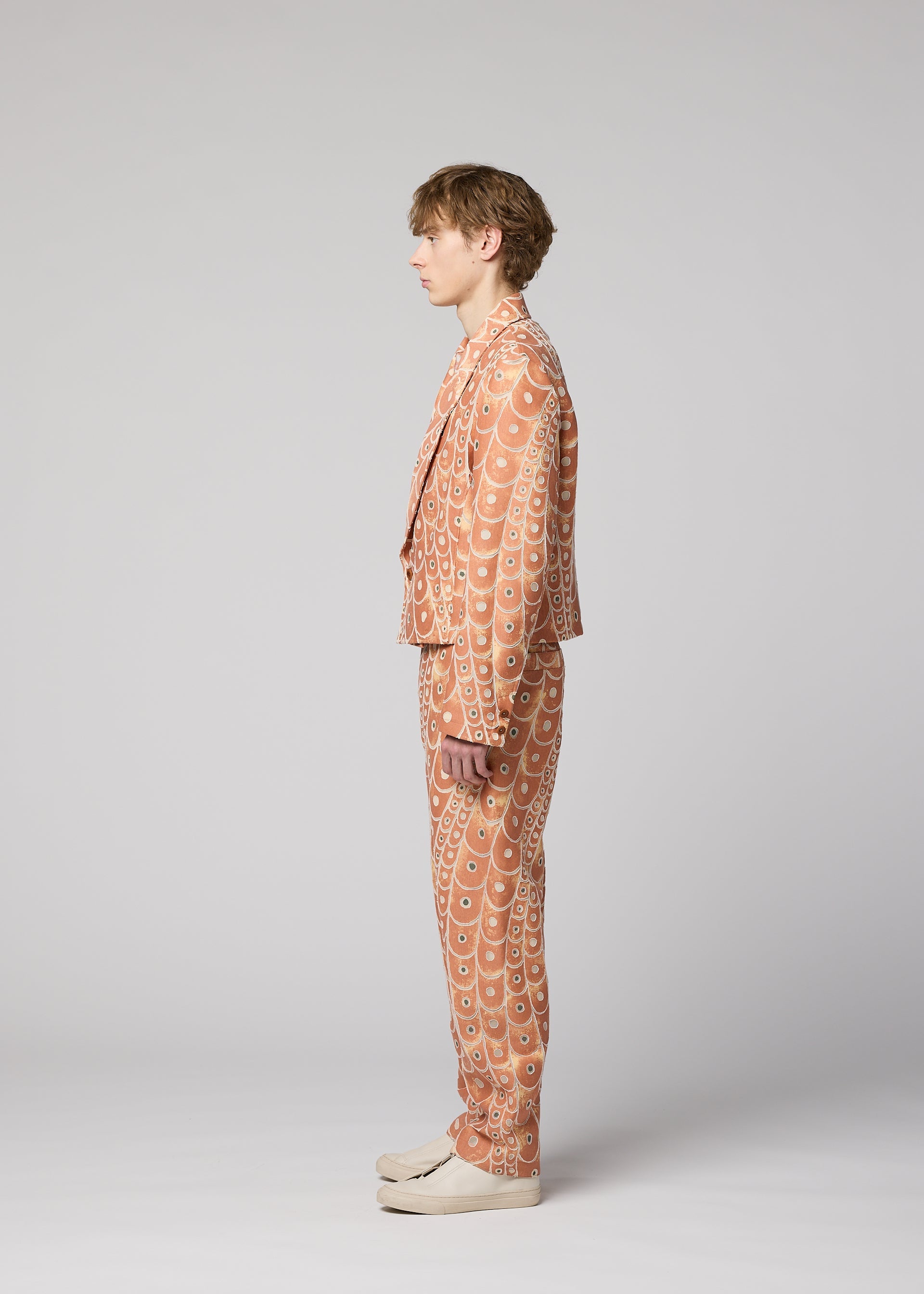 Zoomed view A person stands in profile against a plain light gray background, wearing IM MEN's UROKOMON tan suit with a scale-inspired design by Shoji Kamoda, featuring a short jacket and paired with cream-colored shoes.