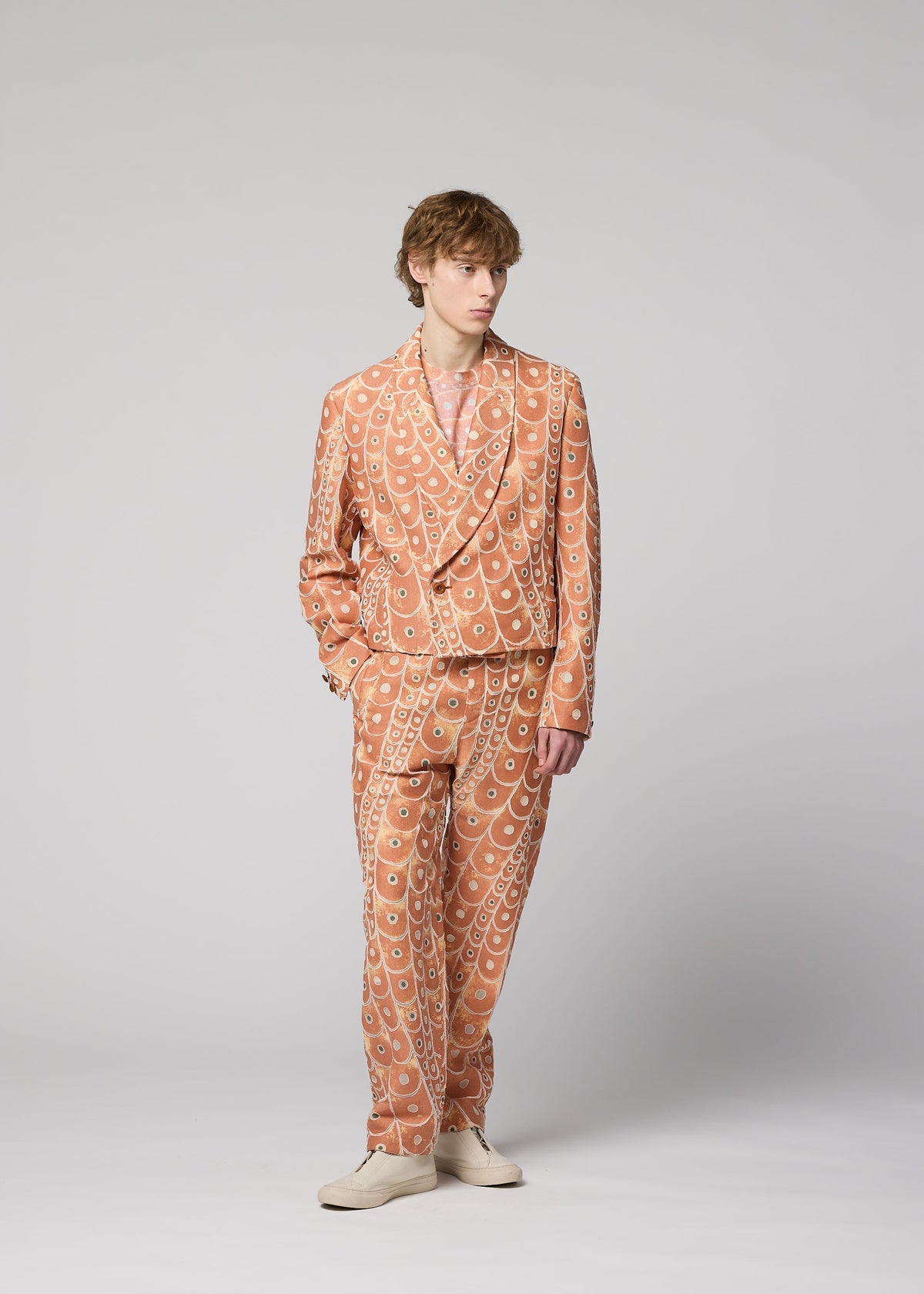 A person with short, wavy hair and a relaxed posture stands against a plain light gray background, wearing the peach-colored UROKOMON jacket and suit by IM MEN, featuring a bold scale-inspired design, paired with light shoes. view 8 of 8