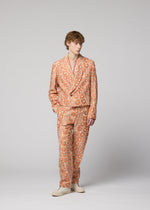A person with short, wavy hair and a relaxed posture stands against a plain light gray background, wearing the peach-colored UROKOMON jacket and suit by IM MEN, featuring a bold scale-inspired design, paired with light shoes.