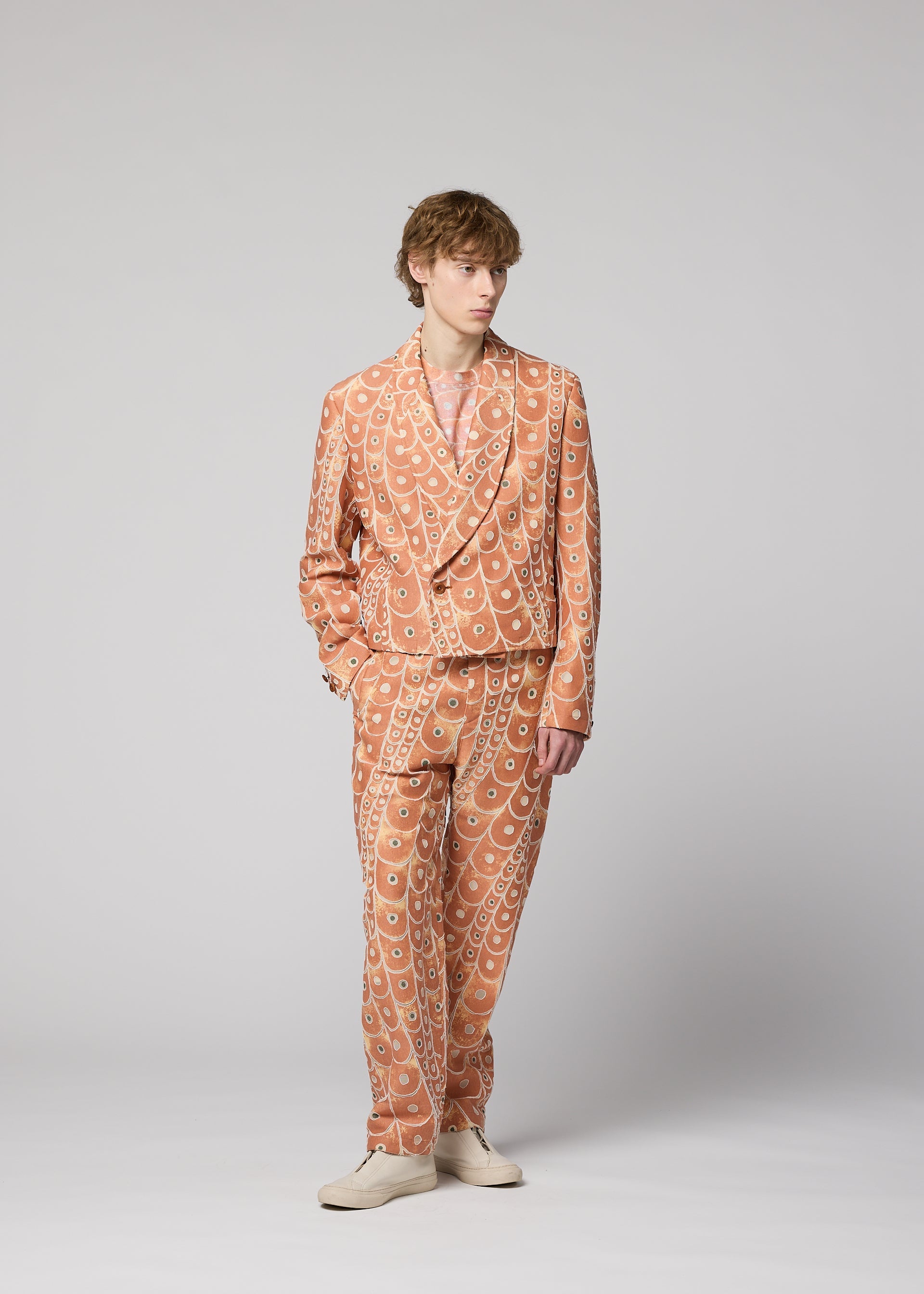 Zoomed view A person with short, wavy hair and a relaxed posture stands against a plain light gray background, wearing the peach-colored UROKOMON jacket and suit by IM MEN, featuring a bold scale-inspired design, paired with light shoes.