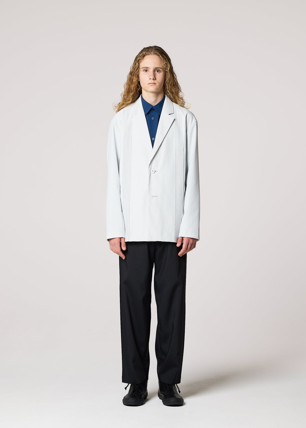 A person with long curly hair faces forward, wearing the IM MEN AIR PLEATS wrinkle-resistant light-colored blazer over a blue shirt, black pants, and black shoes, standing against a plain light background. view 2 of 5
