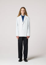 A person with long curly hair faces forward, wearing the IM MEN AIR PLEATS wrinkle-resistant light-colored blazer over a blue shirt, black pants, and black shoes, standing against a plain light background.