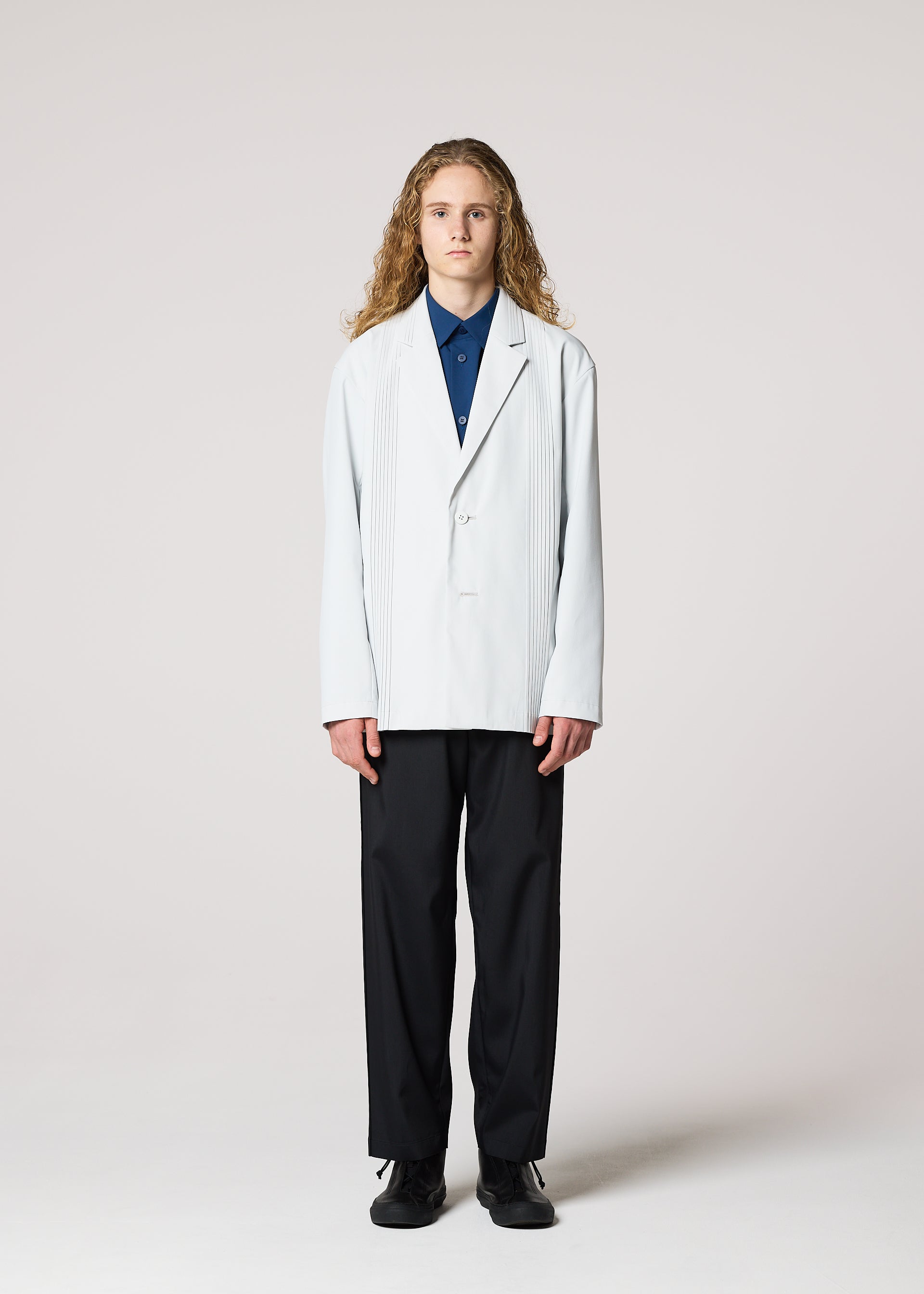 Zoomed view A person with long curly hair faces forward, wearing the IM MEN AIR PLEATS wrinkle-resistant light-colored blazer over a blue shirt, black pants, and black shoes, standing against a plain light background.