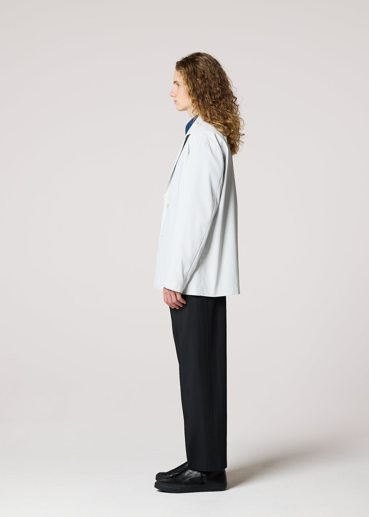 A person with long curly hair stands in profile against a plain light background, wearing IM MEN's AIR PLEATS wrinkle-resistant tailored jacket, dark trousers, and black shoes. view 3 of 5