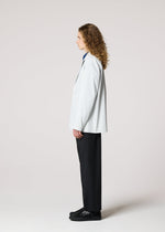 A person with long curly hair stands in profile against a plain light background, wearing IM MEN's AIR PLEATS wrinkle-resistant tailored jacket, dark trousers, and black shoes.