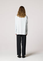 A person with curly hair stands facing away, wearing IM MEN's AIR PLEATS light gray wrinkle-resistant long-sleeve shirt, black pants, and black shoes against a plain light background.