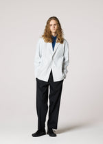 A person with long curly hair stands against a plain background, wearing IM MEN's AIR PLEATS white textured blazer over a blue shirt, black wide-leg pants, and black shoes, hands in pockets and a neutral expression.
