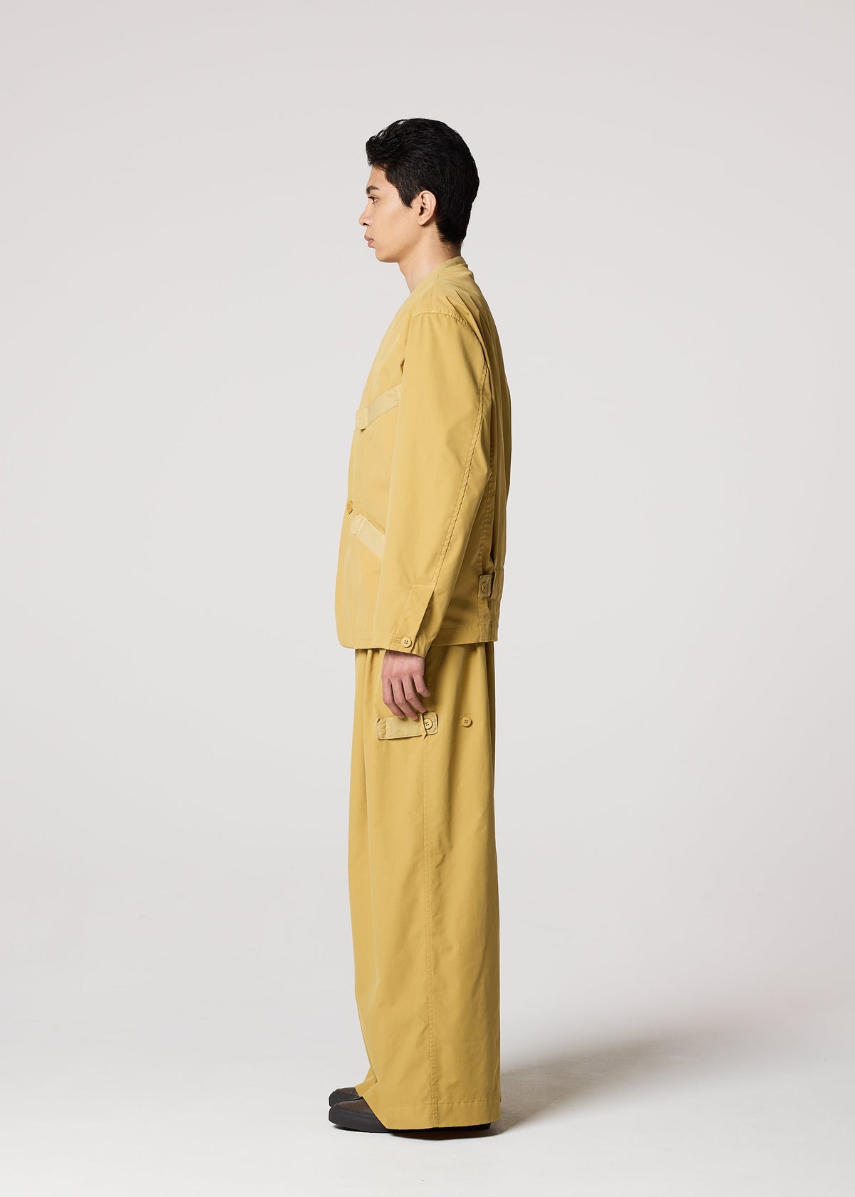 A person stands in profile against a plain light background, wearing IM MEN's RESILIENCE set—wrinkle-resistant, lightweight polyester jacket and matching wide-leg pants with patch pockets—styled with dark shoes for comfort and style. view 7 of 10