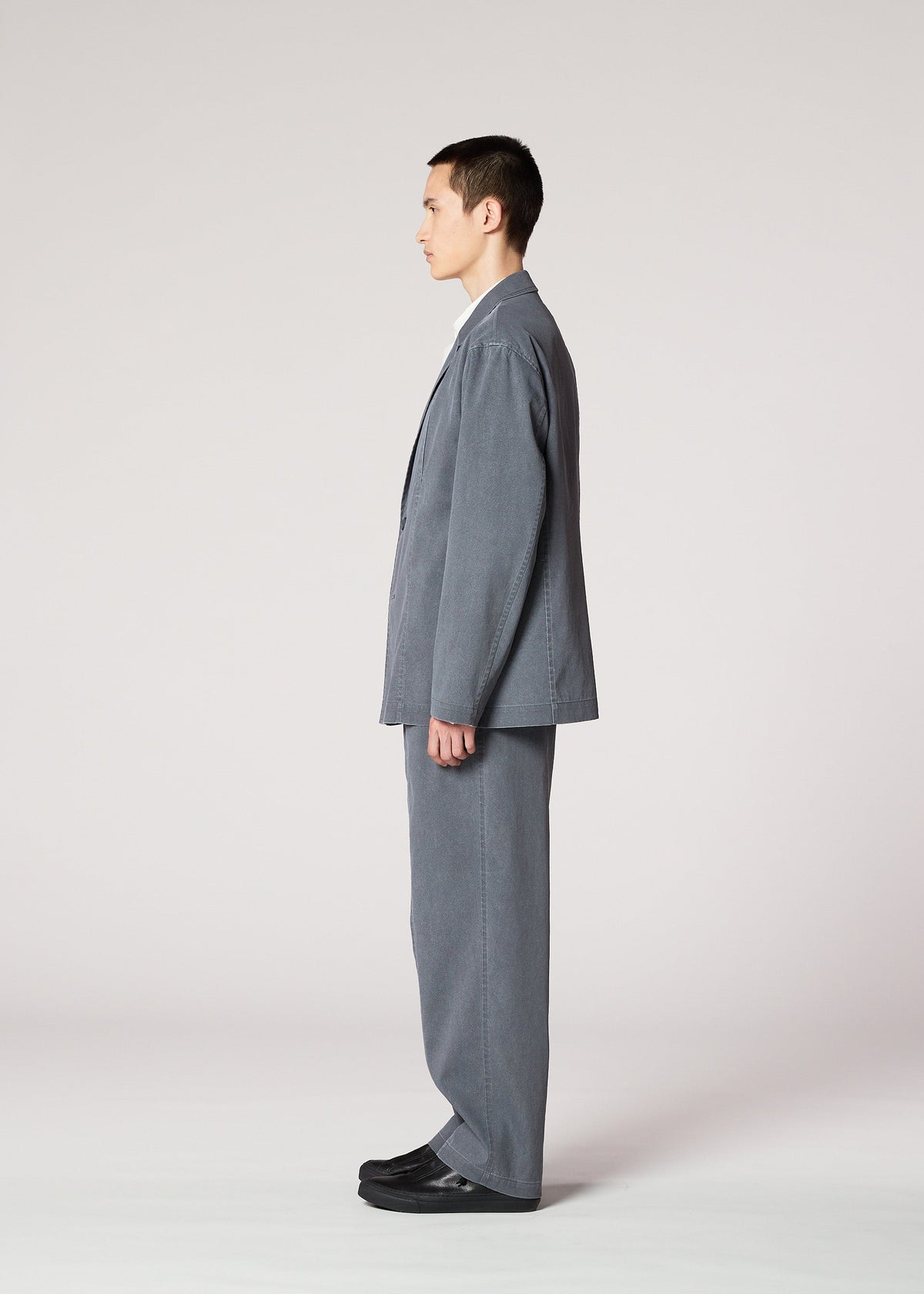 A man in profile faces left, wearing IM MEN's AIR WASH loose gray suit made from plant-derived polyester with wide pants and black shoes, set against a plain light background. view 6 of 8