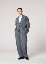 A person wears the IM MEN AIR WASH oversized gray suit in plant-derived polyester, paired with wide-leg pants, a white shirt, and black shoes. Hands are in pockets, standing against a plain background with a neutral expression.