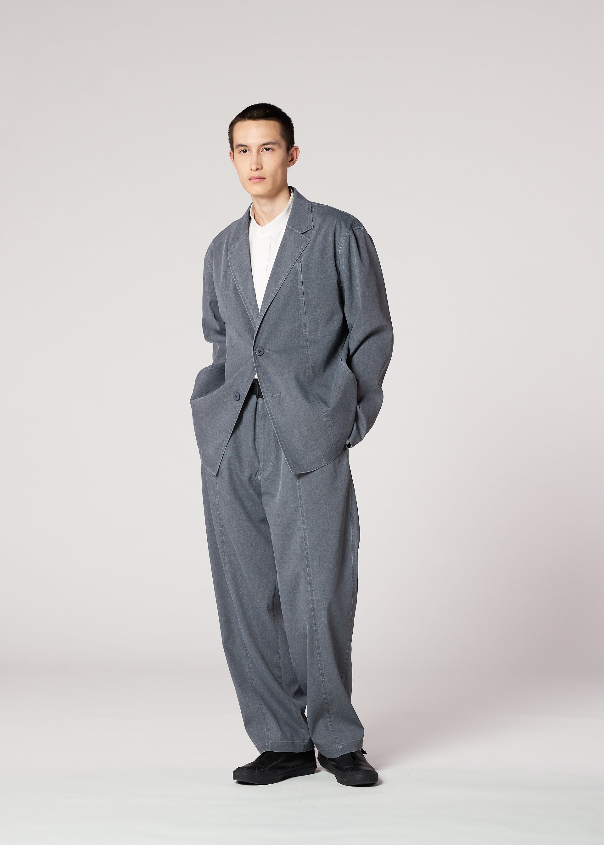 Zoomed view A person wears the IM MEN AIR WASH oversized gray suit in plant-derived polyester, paired with wide-leg pants, a white shirt, and black shoes. Hands are in pockets, standing against a plain background with a neutral expression.