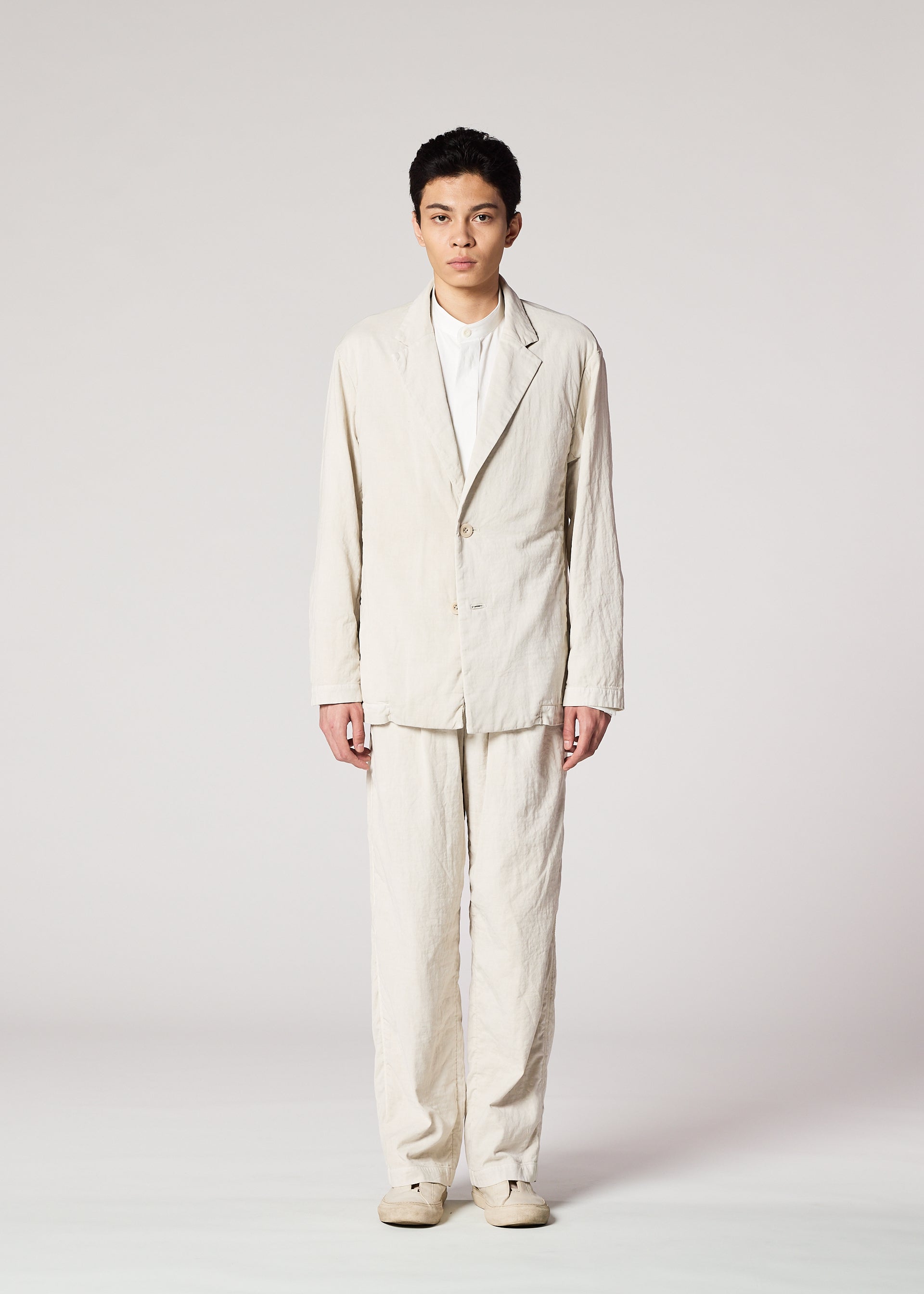 Zoomed view A person faces forward against a plain background, wearing the IM MEN PIGMENT DOUBLE FACE COTTON suit: a light, pigment-washed single-button jacket with matching pants, white shirt underneath, and light shoes.