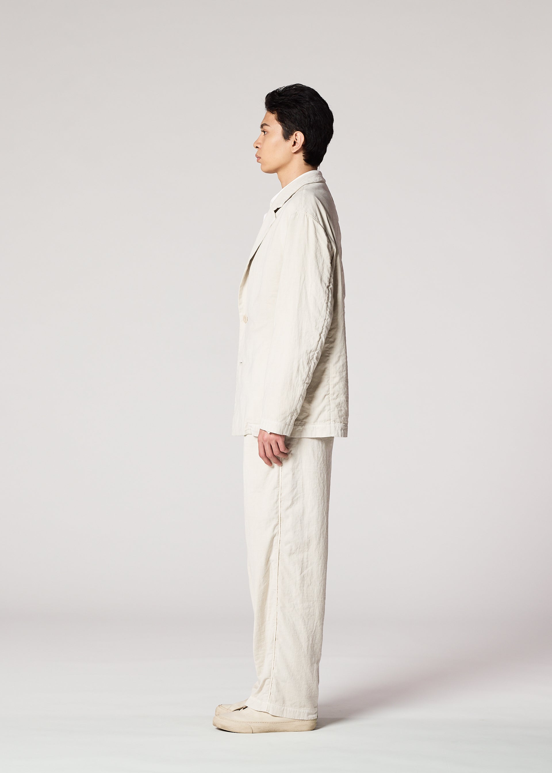 Zoomed view A person in profile stands straight against a plain light background, dressed in IM MEN's PIGMENT DOUBLE FACE COTTON off-white suit with matching shoes, hands relaxed at their sides.