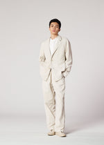 Against a plain, light background, a person wears IM MEN's PIGMENT DOUBLE FACE COTTON suit—loose-fitting with a soft-textured jacket—paired with a white shirt and matching shoes, hands in pockets, looking confidently at the camera.