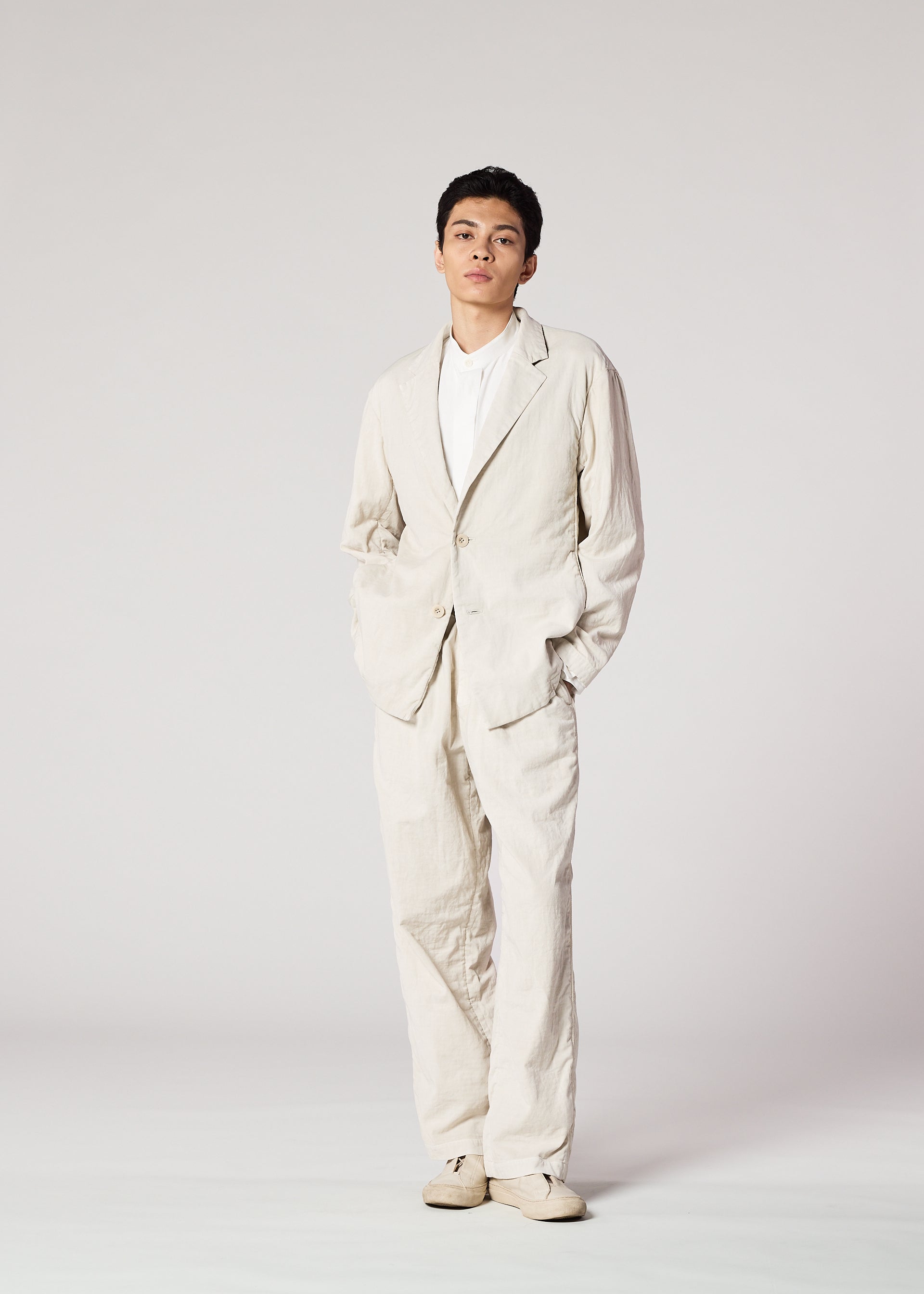 Zoomed view Against a plain, light background, a person wears IM MEN's PIGMENT DOUBLE FACE COTTON suit—loose-fitting with a soft-textured jacket—paired with a white shirt and matching shoes, hands in pockets, looking confidently at the camera.