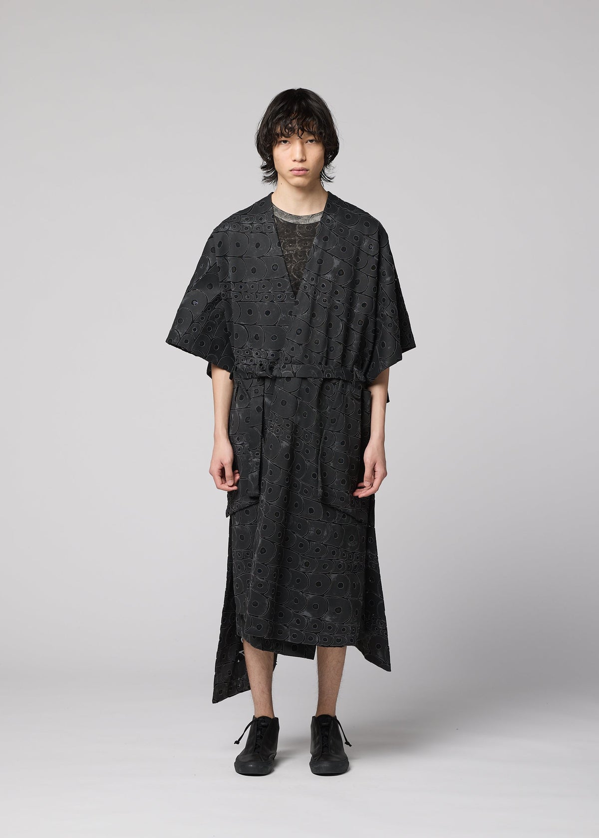 A person with medium-length dark hair faces forward, wearing the IM MEN UROKOMON kimono-style robe in black with a subtle scale pattern, short sleeves, knee length, and a belt. Black shoes complete the look against a plain light grey background. view 6 of 9
