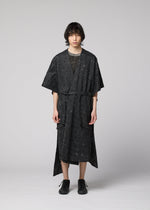 A person with medium-length dark hair faces forward, wearing the IM MEN UROKOMON kimono-style robe in black with a subtle scale pattern, short sleeves, knee length, and a belt. Black shoes complete the look against a plain light grey background.
