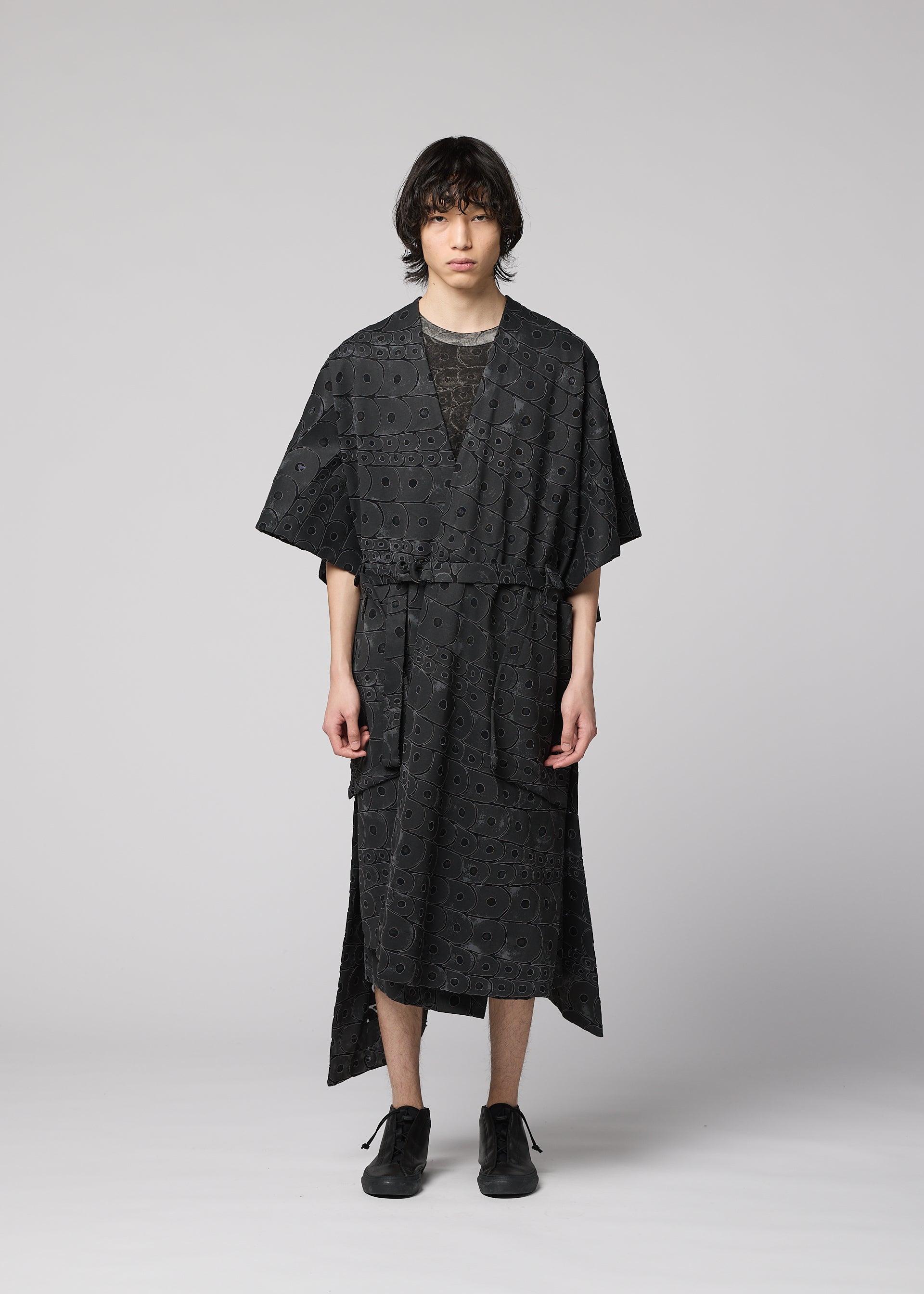 Zoomed view A person with medium-length dark hair faces forward, wearing the IM MEN UROKOMON kimono-style robe in black with a subtle scale pattern, short sleeves, knee length, and a belt. Black shoes complete the look against a plain light grey background.
