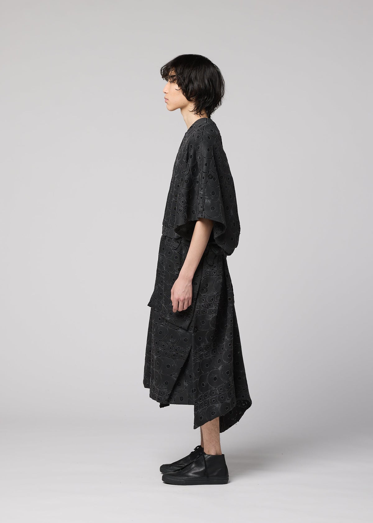 A person with medium-length dark hair stands in profile against a light background, wearing IM MEN’s UROKOMON vest—a ceramic-inspired piece with a subtle scale design—over a loose black eyelet dress and black shoes. view 7 of 9