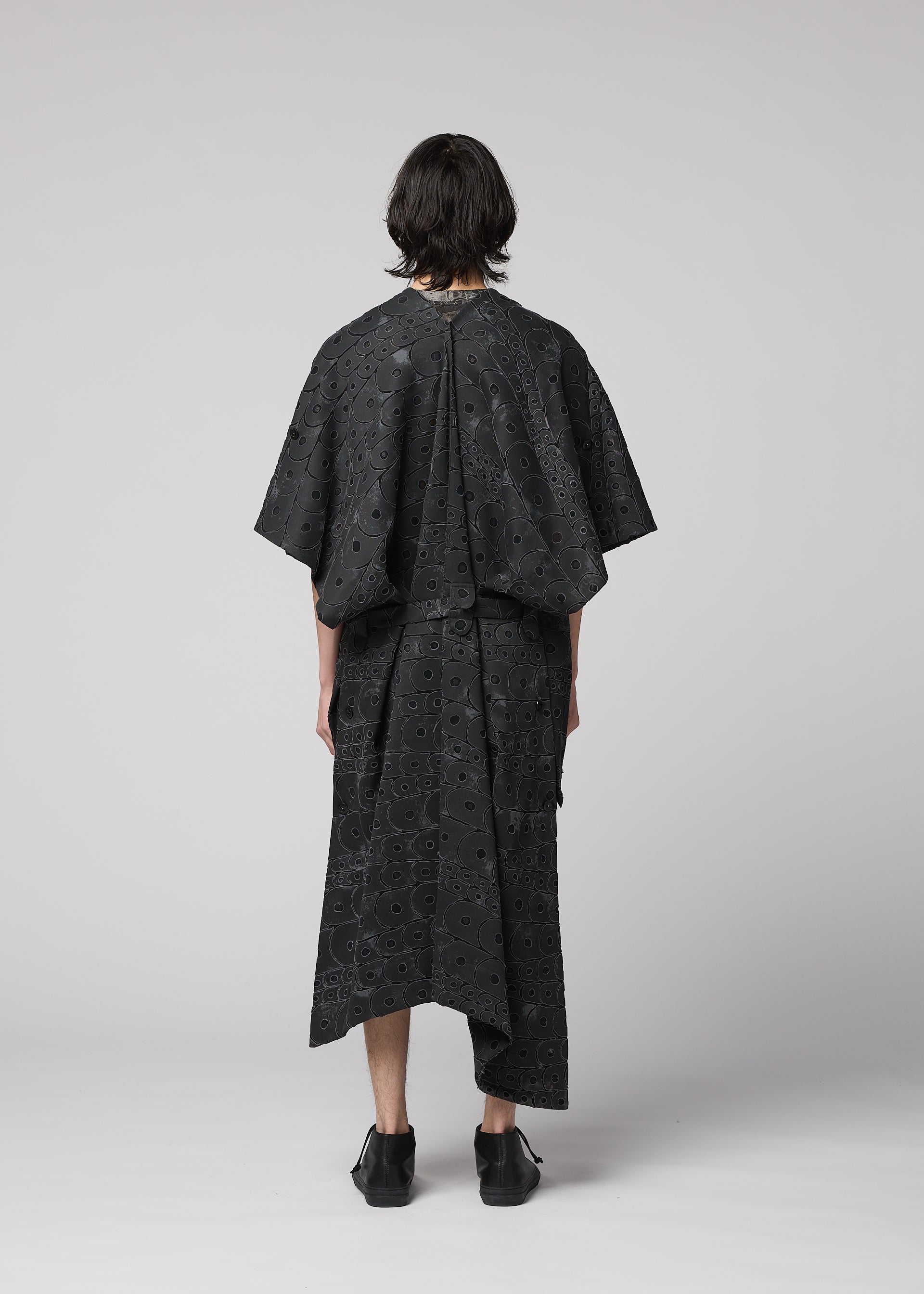 Zoomed view A person stands with their back to the camera, wearing IM MEN's UROKOMON: a loose black outfit with a scale-inspired ceramic vest, eyelet patterns, wide sleeves, an asymmetrical skirt, and black shoes against a light gray background.