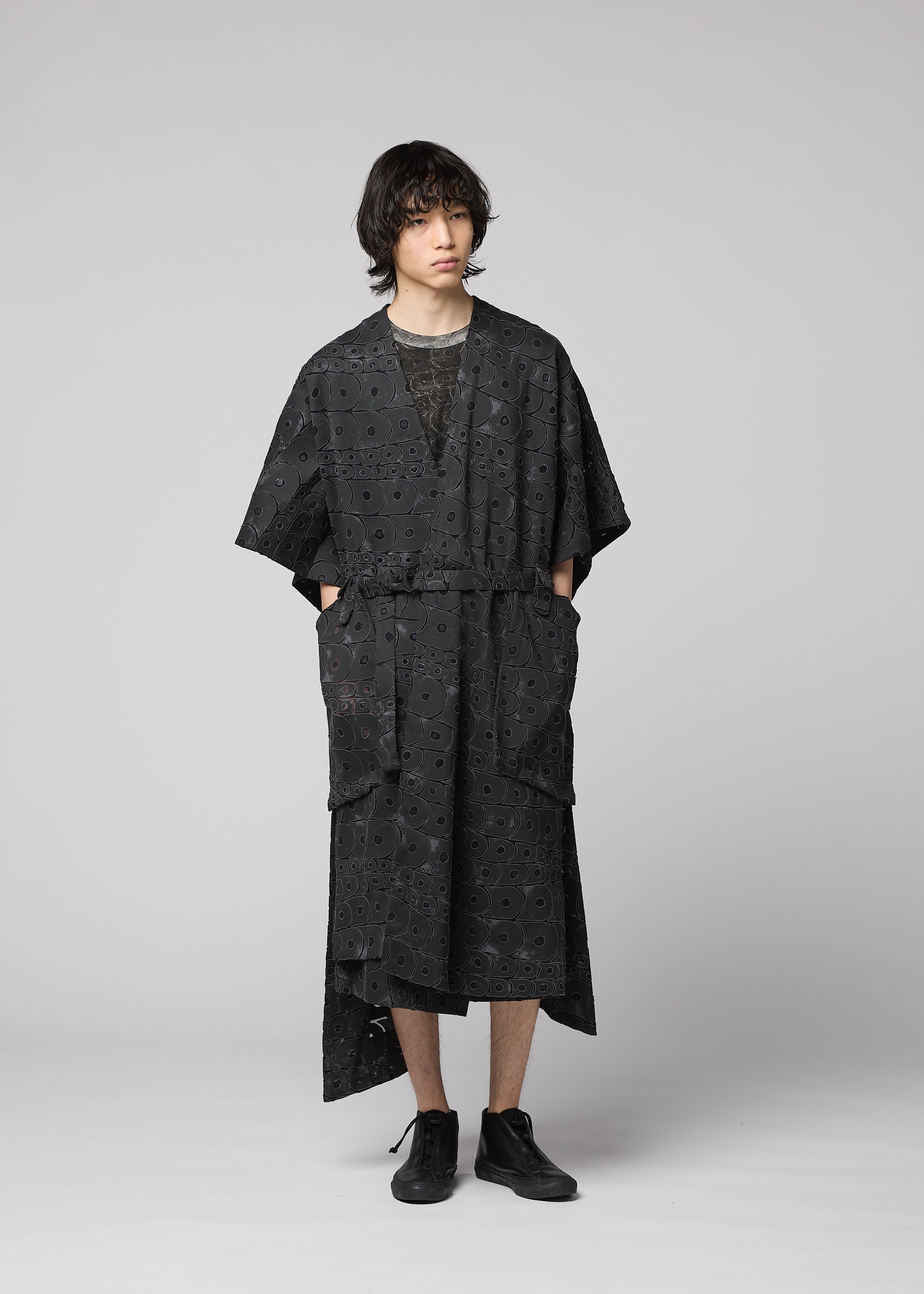 Zoomed view A person with wavy dark hair wears the IM MEN UROKOMON, a loose black kimono-style outfit with wide sleeves and large front pockets, styled with black shoes and a ceramic-inspired vest, standing against a plain light gray background.