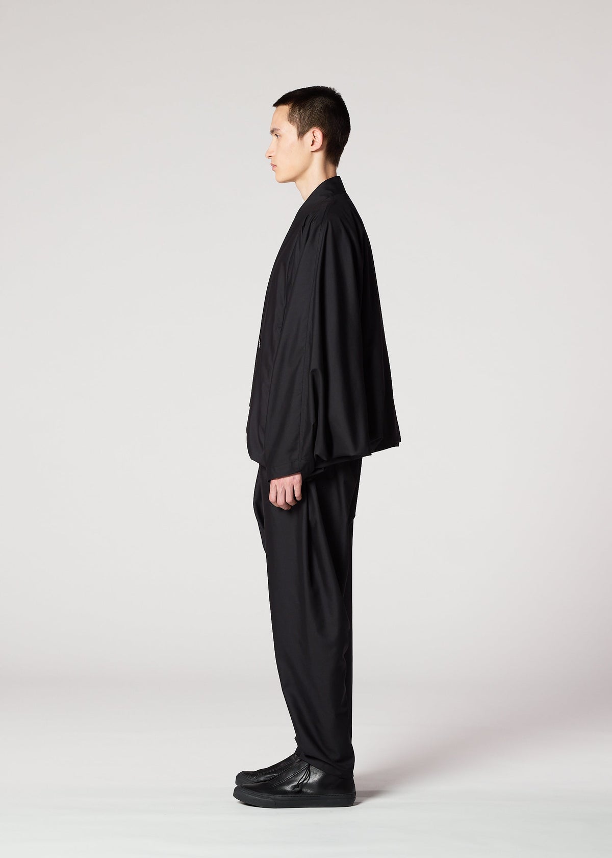 A person with short dark hair stands in profile, wearing the IM MEN SWING BLACK suit—a loose black jacket and wrinkle-resistant pants with a loose silhouette—paired with black shoes, set against a plain light gray background. view 4 of 7