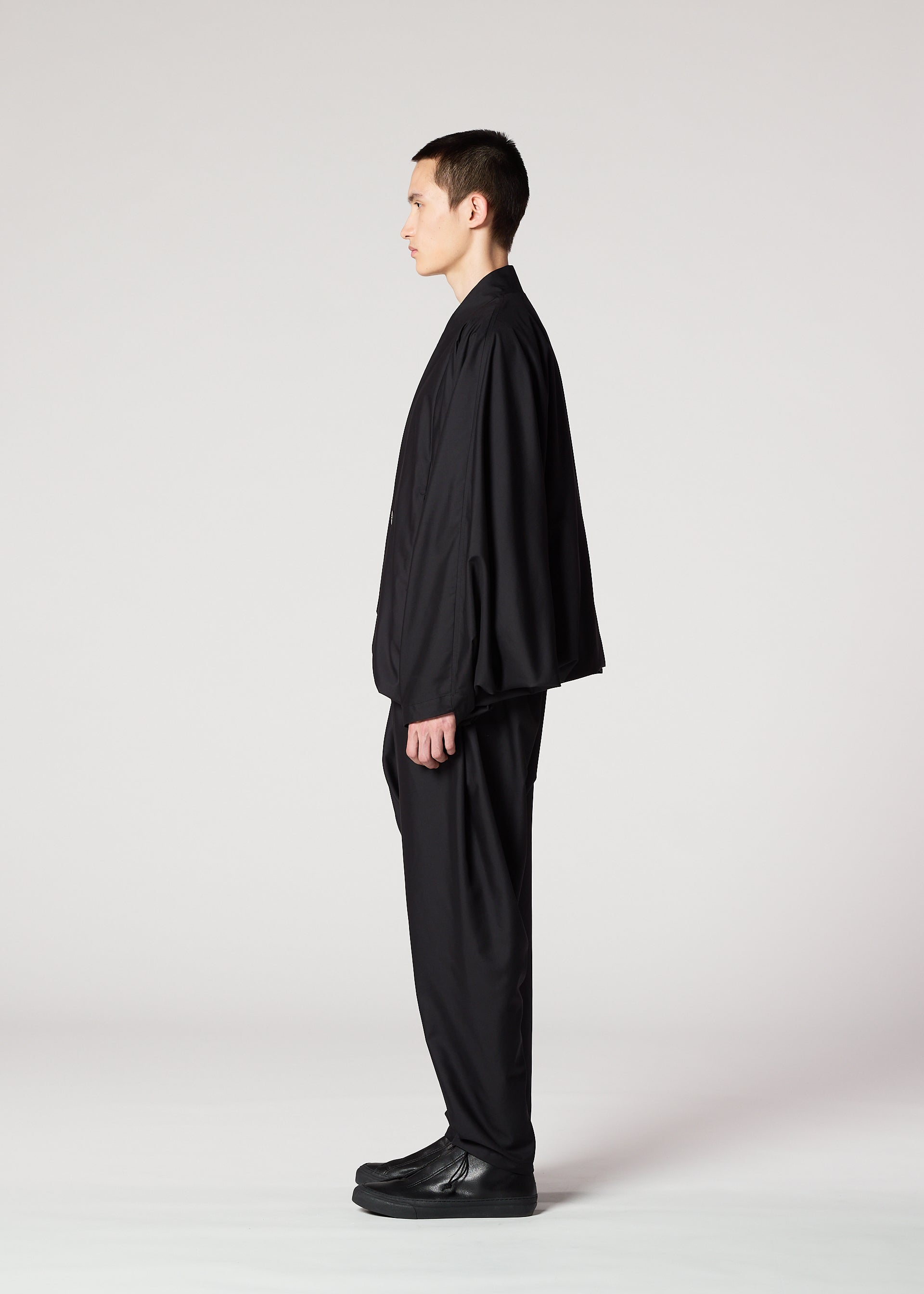 Zoomed view A person with short dark hair stands in profile, wearing the IM MEN SWING BLACK suit—a loose black jacket and wrinkle-resistant pants with a loose silhouette—paired with black shoes, set against a plain light gray background.