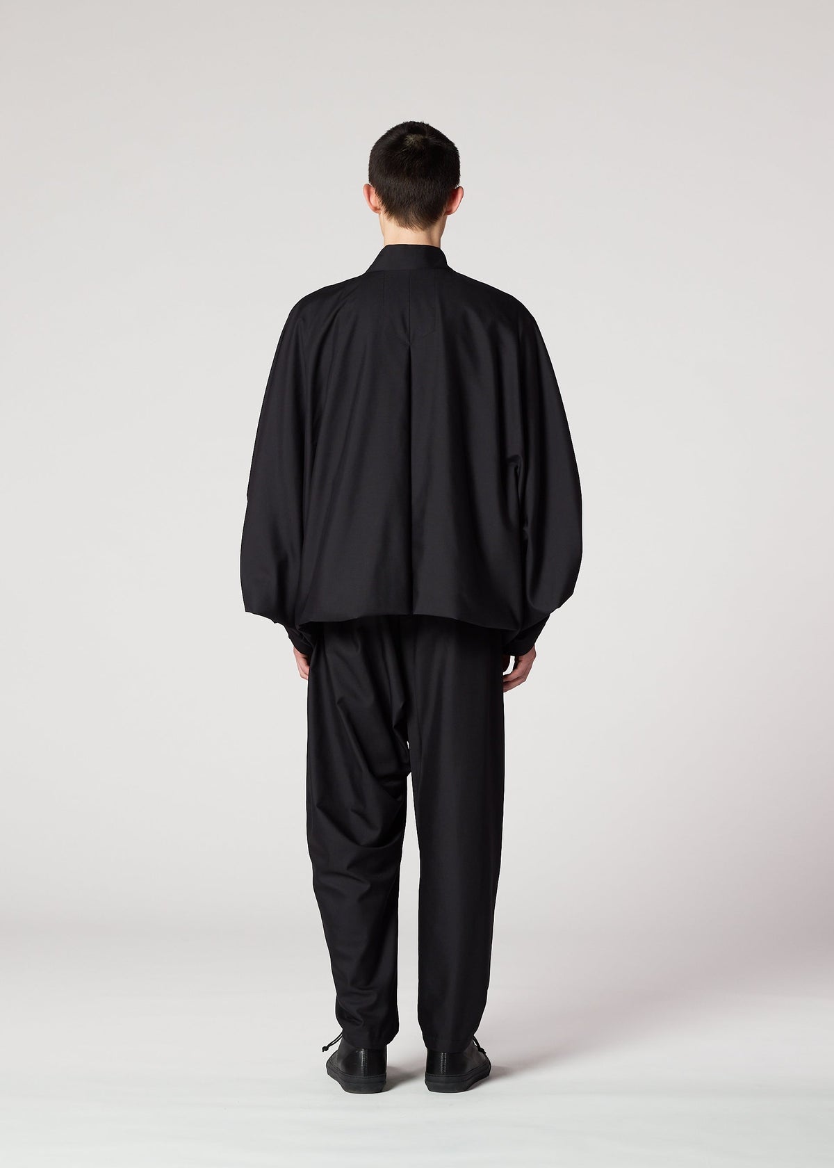 A person stands facing away from the camera, wearing IM MEN’s SWING BLACK wrinkle-resistant pants and an oversized jacket with a loose silhouette, set against a plain light gray background. view 6 of 7