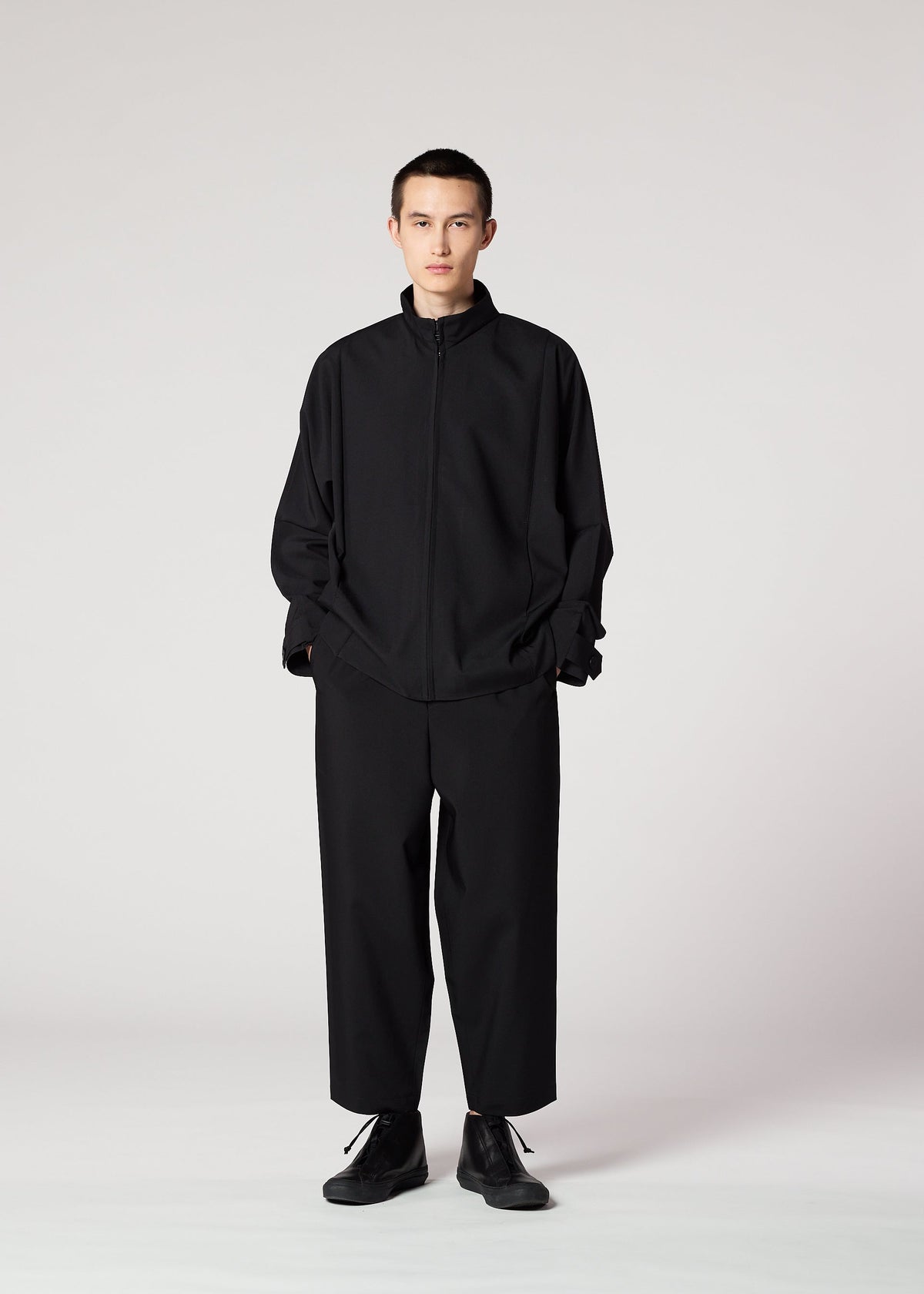 A person stands against a light background, wearing the IM MEN FLAT BLACK zip-up jacket and wide-leg, wrinkle-resistant pants, hands in pockets and a neutral expression. view 8 of 8
