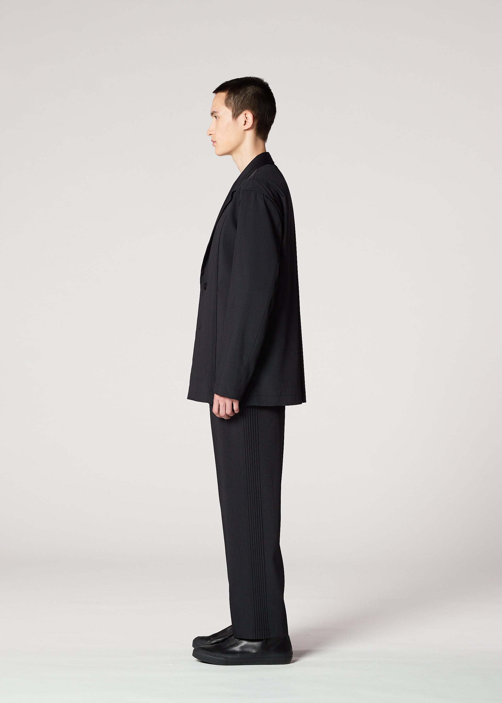 Zoomed view A person with short dark hair is seen in profile wearing IM MEN's AIR PLEATS BLACK suit and lightweight pants with black shoes, standing against a plain light background.