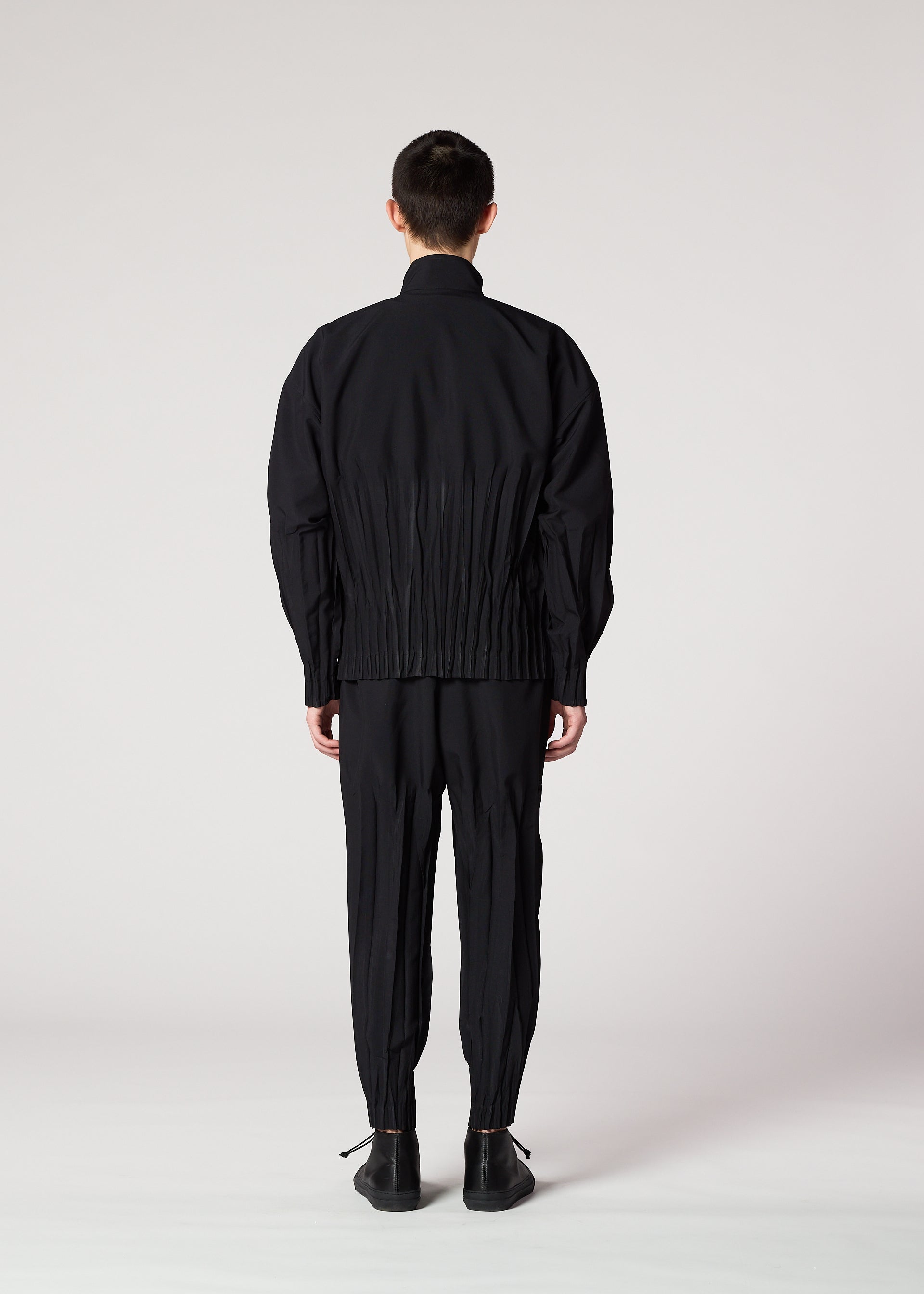 Zoomed view A person with short dark hair faces away, wearing the IM MEN WAFT BLACK wrinkle-resistant pants with gathered details and a loose black jacket, paired with black shoes against a plain light background.