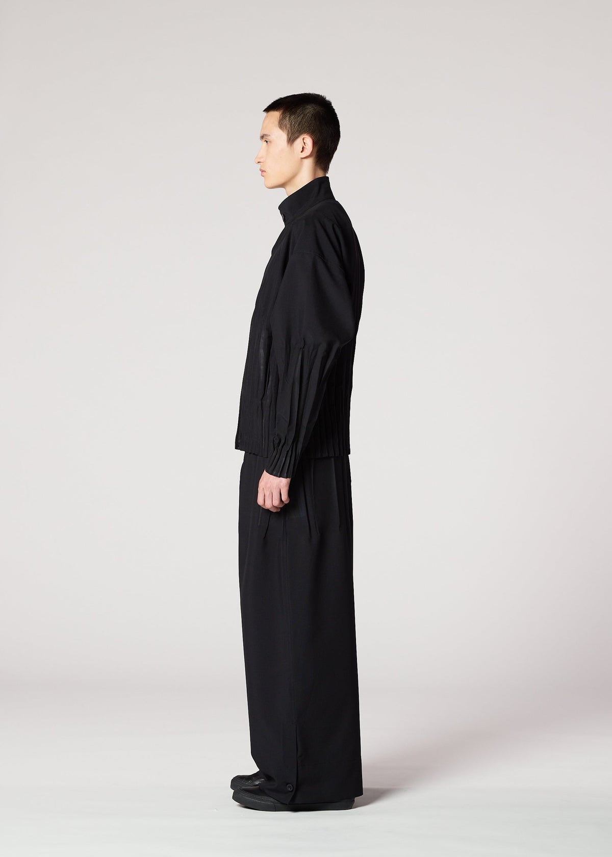 A person in profile stands against a plain light background, dressed in IM MEN's WAFT BLACK: a loose-fitting black jacket and wide, wrinkle-resistant pants paired with black shoes for a minimalist, modern look. view 11 of 15