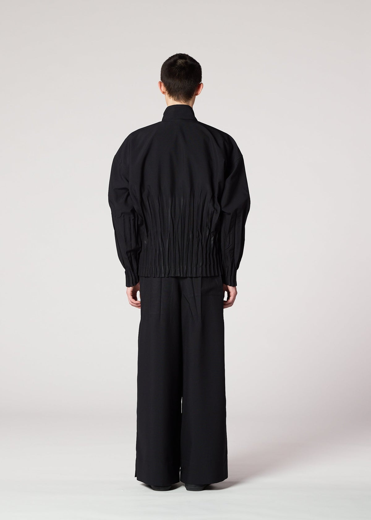 A person with short hair faces away, wearing IM MEN's WAFT BLACK— a loose black jacket with pleats and wide, linen-textured pants— against a light background. view 6 of 7