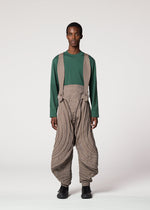 A person stands facing forward in IM MEN’s ENGRAVE look: a green long-sleeve shirt with Shoji Kamoda inspired loose tan pants featuring curved lines, matching suspenders, black shoes, and short hair against a plain background.