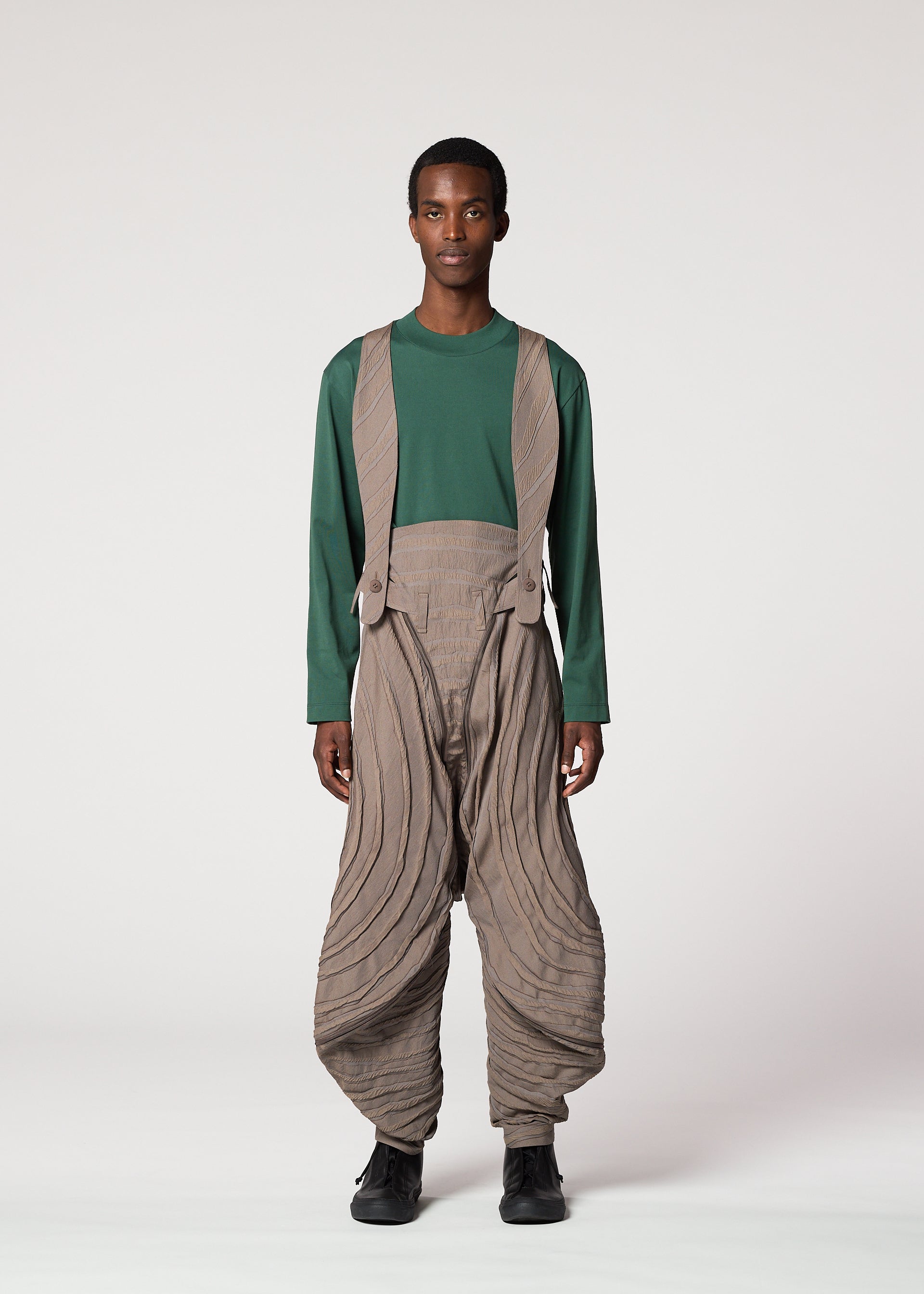Zoomed view A person stands facing forward in IM MEN’s ENGRAVE look: a green long-sleeve shirt with Shoji Kamoda inspired loose tan pants featuring curved lines, matching suspenders, black shoes, and short hair against a plain background.