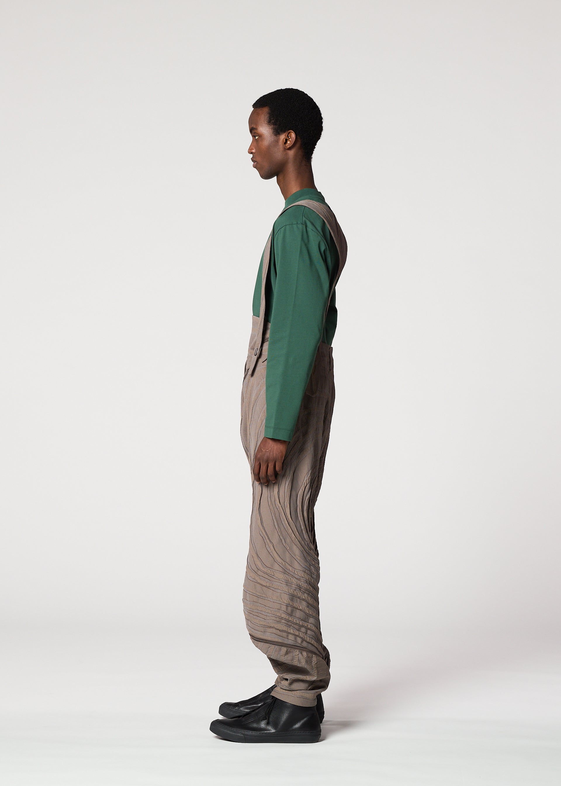 Zoomed view A person stands in profile against a white background, wearing the ENGRAVE long-sleeve shirt and Shoji Kamoda inspired textured tan overalls by IM MEN, paired with black shoes.