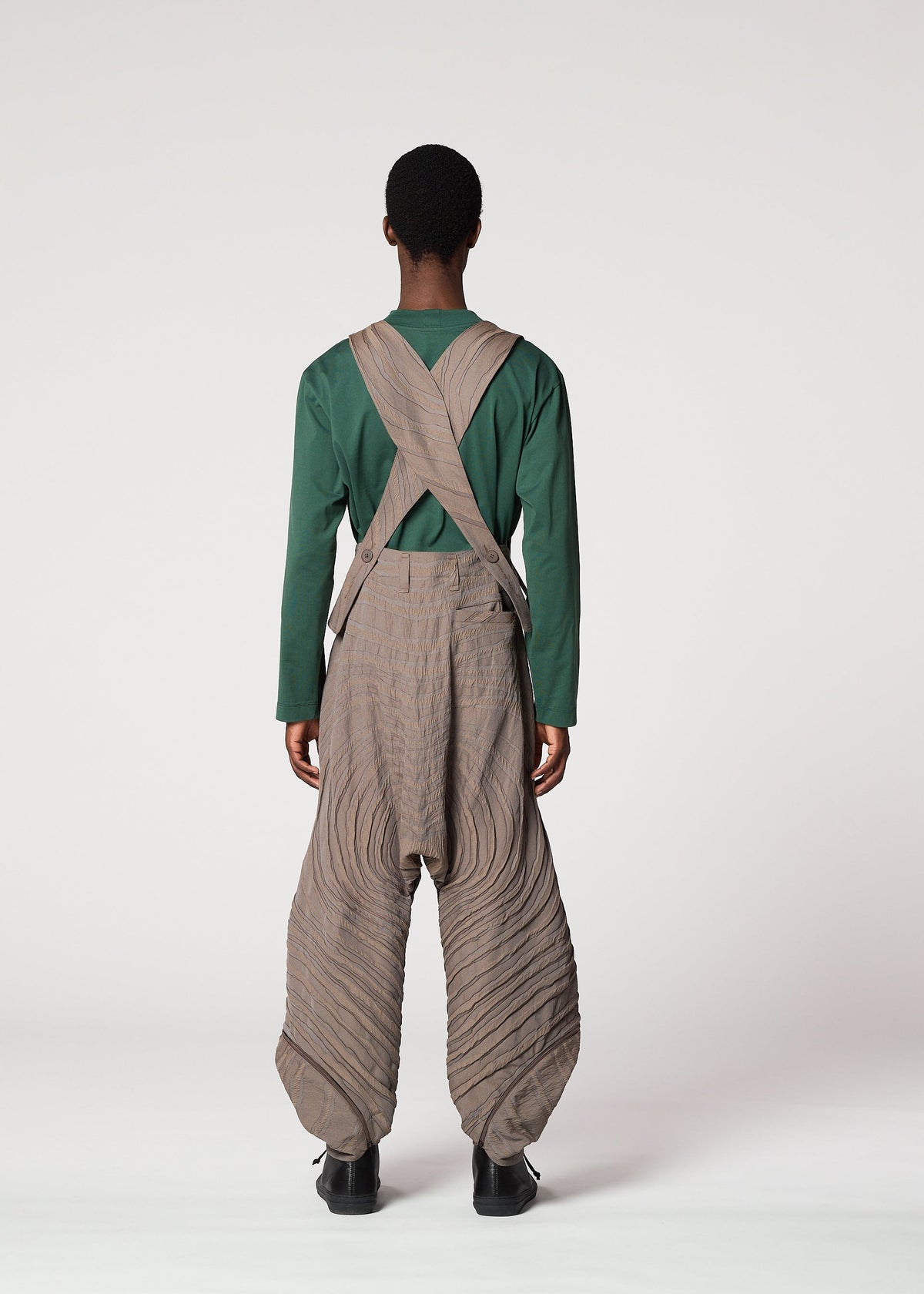 A person stands facing away, wearing IM MEN's ENGRAVE—green long-sleeve shirt and taupe overalls with crossed straps and wide, ridged legs. The background is plain white. view 7 of 8