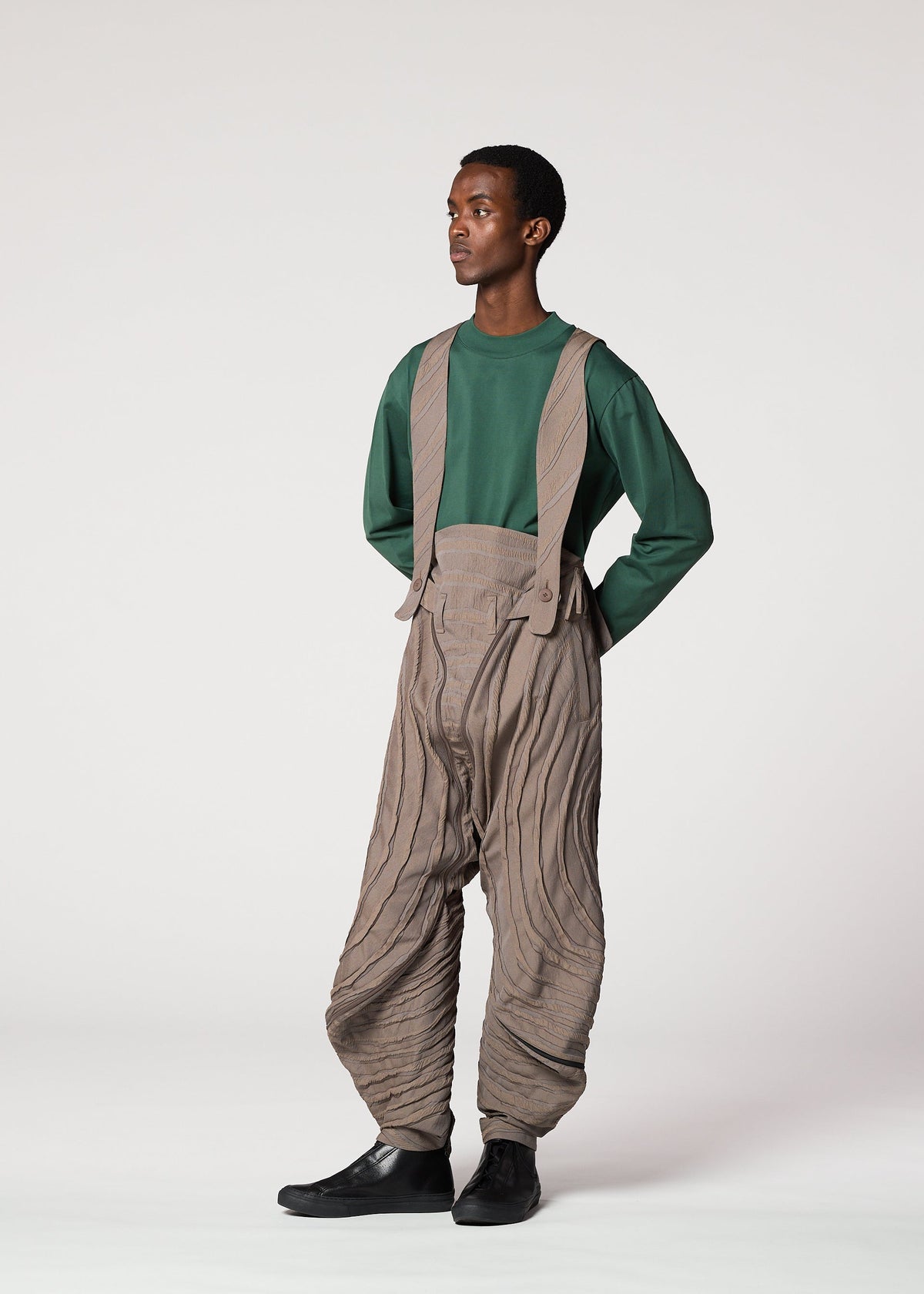 A person with short hair and black shoes stands against a plain background wearing IM MEN’s ENGRAVE: green long-sleeve top and high-waisted Shoji Kamoda–inspired pants with suspenders and textured wavy patterns. view 8 of 8