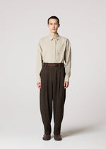 A person faces forward against a plain light background, wearing a beige long-sleeve shirt tucked into high-waisted TEXTURES pants by IM MEN, inspired by Shoji Kamoda ceramics, with a belt and brown shoes.