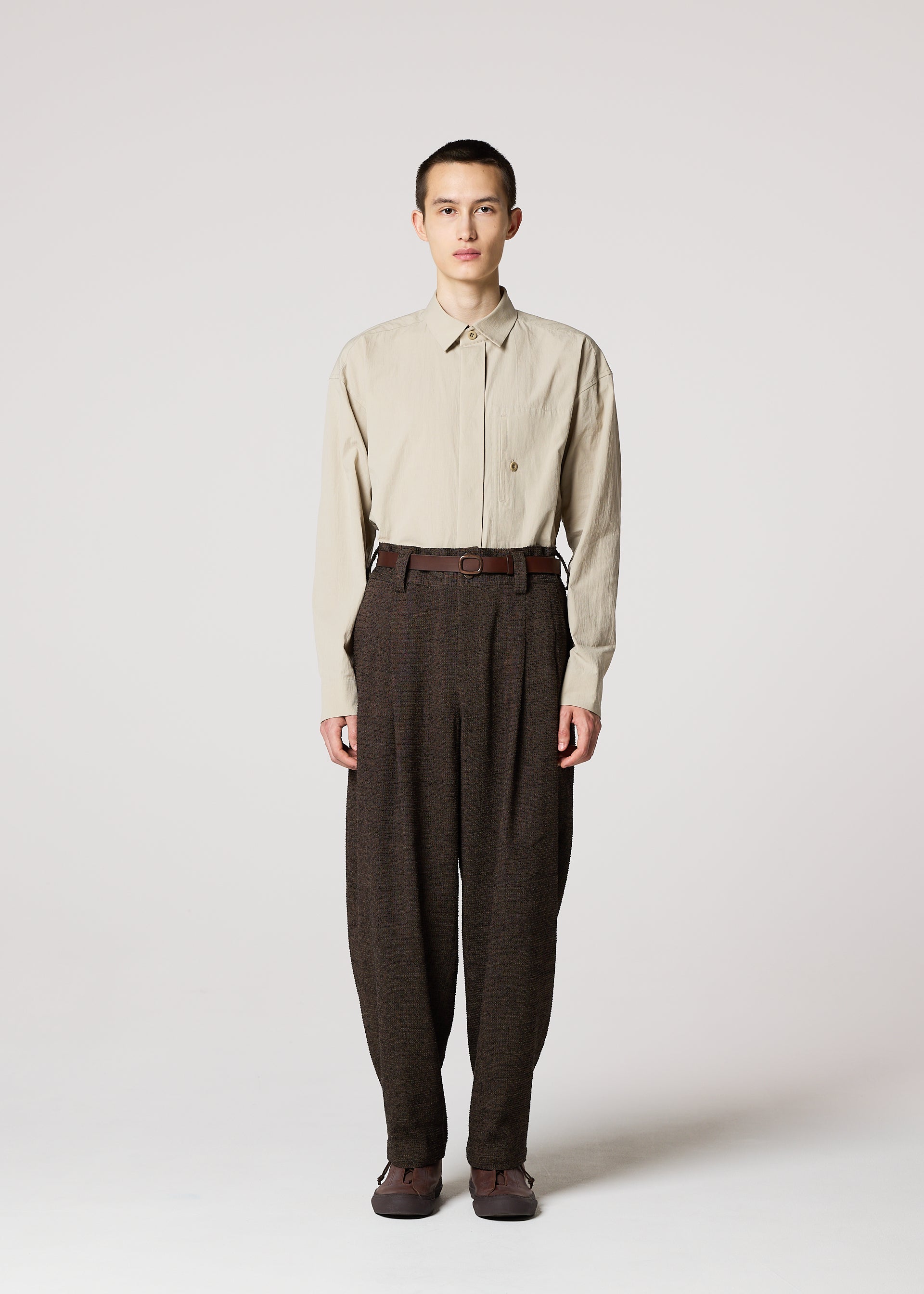 Zoomed view A person faces forward against a plain light background, wearing a beige long-sleeve shirt tucked into high-waisted TEXTURES pants by IM MEN, inspired by Shoji Kamoda ceramics, with a belt and brown shoes.