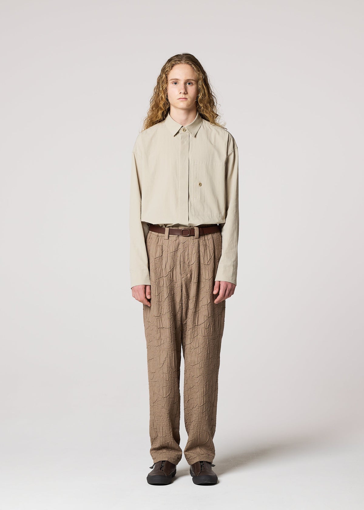 A person with long curly hair faces forward in IM MEN’s SHRINK JACQUARD: a beige button-up shirt tucked into wood-grain woven pants made on a computer jacquard loom, styled with a brown belt and dark shoes against a plain background. view 5 of 8