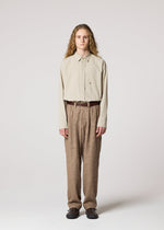 A person with long curly hair faces forward in IM MEN’s SHRINK JACQUARD: a beige button-up shirt tucked into wood-grain woven pants made on a computer jacquard loom, styled with a brown belt and dark shoes against a plain background.
