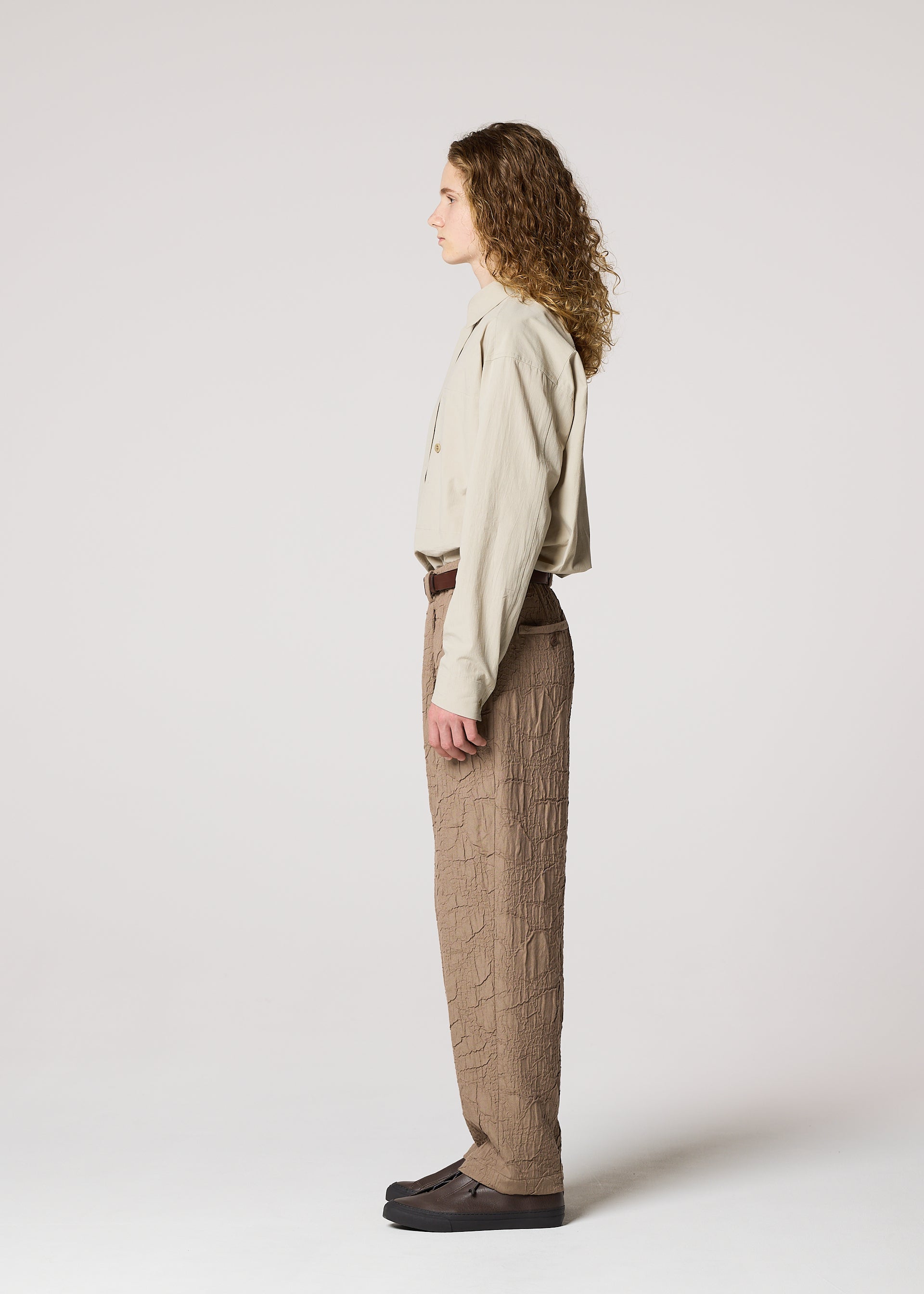Zoomed view A person with long curly hair stands in profile against a light background, wearing IM MEN's SHRINK JACQUARD textured wood-grain woven pants, a beige shirt, and dark shoes.